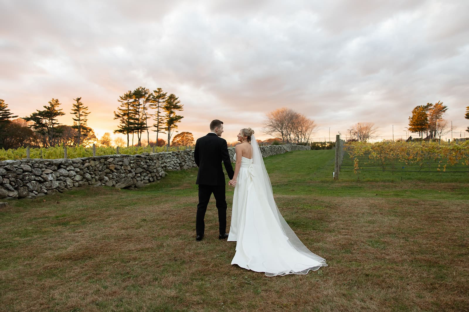 Bride and groom walking together at sunset along a stone wall and vineyard landscape at one of the scenic Wedding Venues in Rhode Island.
