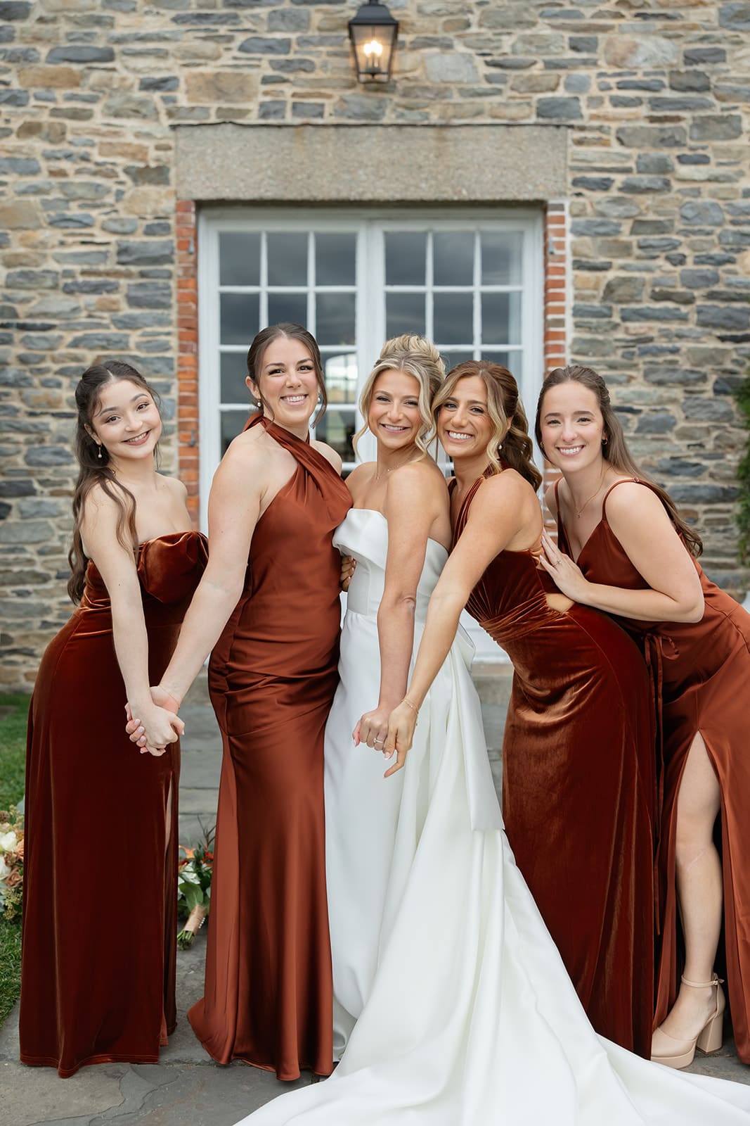 Bride with bridesmaids in rust velvet dresses in front of the stone manor at their Shepherd’s Run wedding.