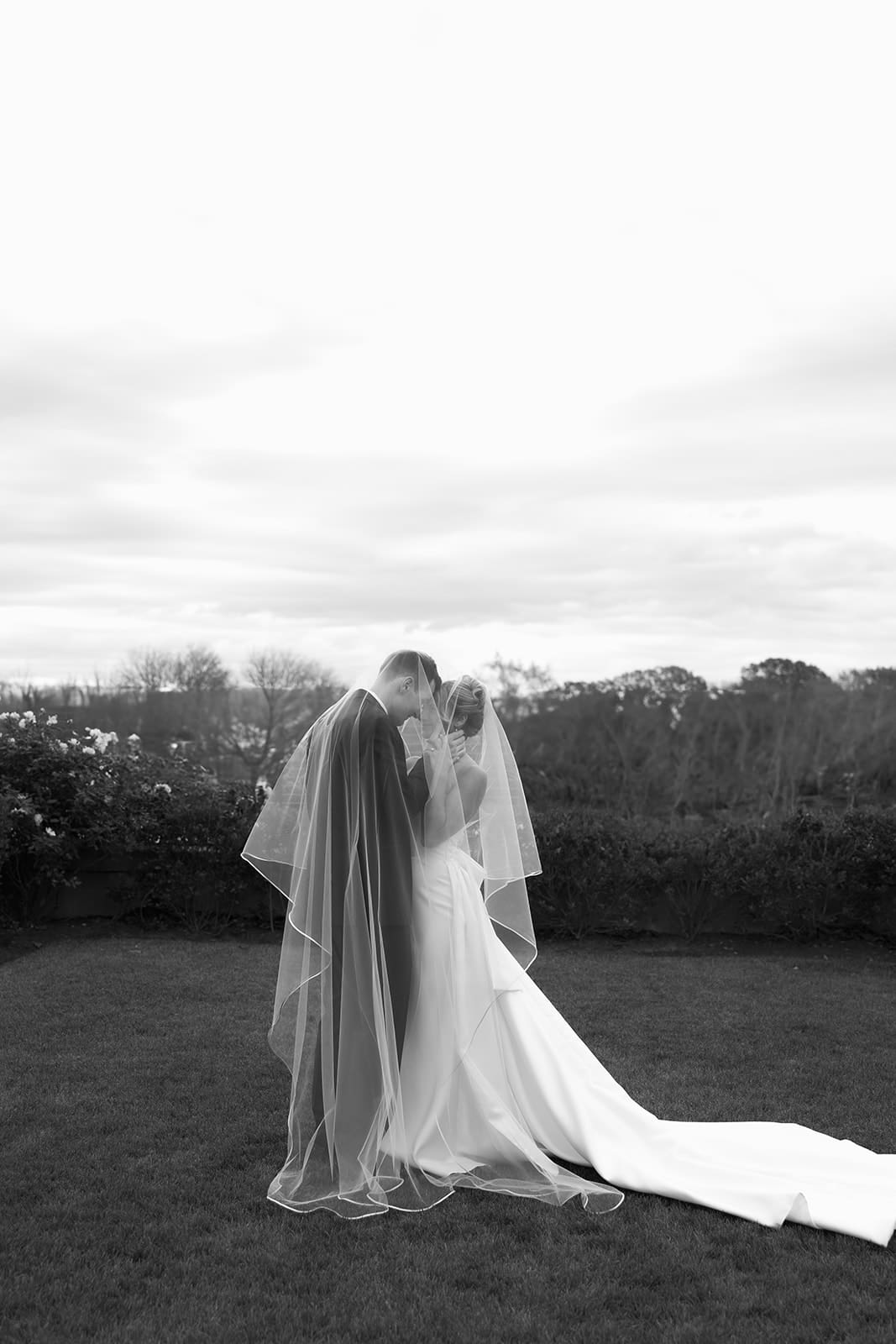 Bride and groom embracing under flowing veil on estate lawn in romantic black and white portrait.