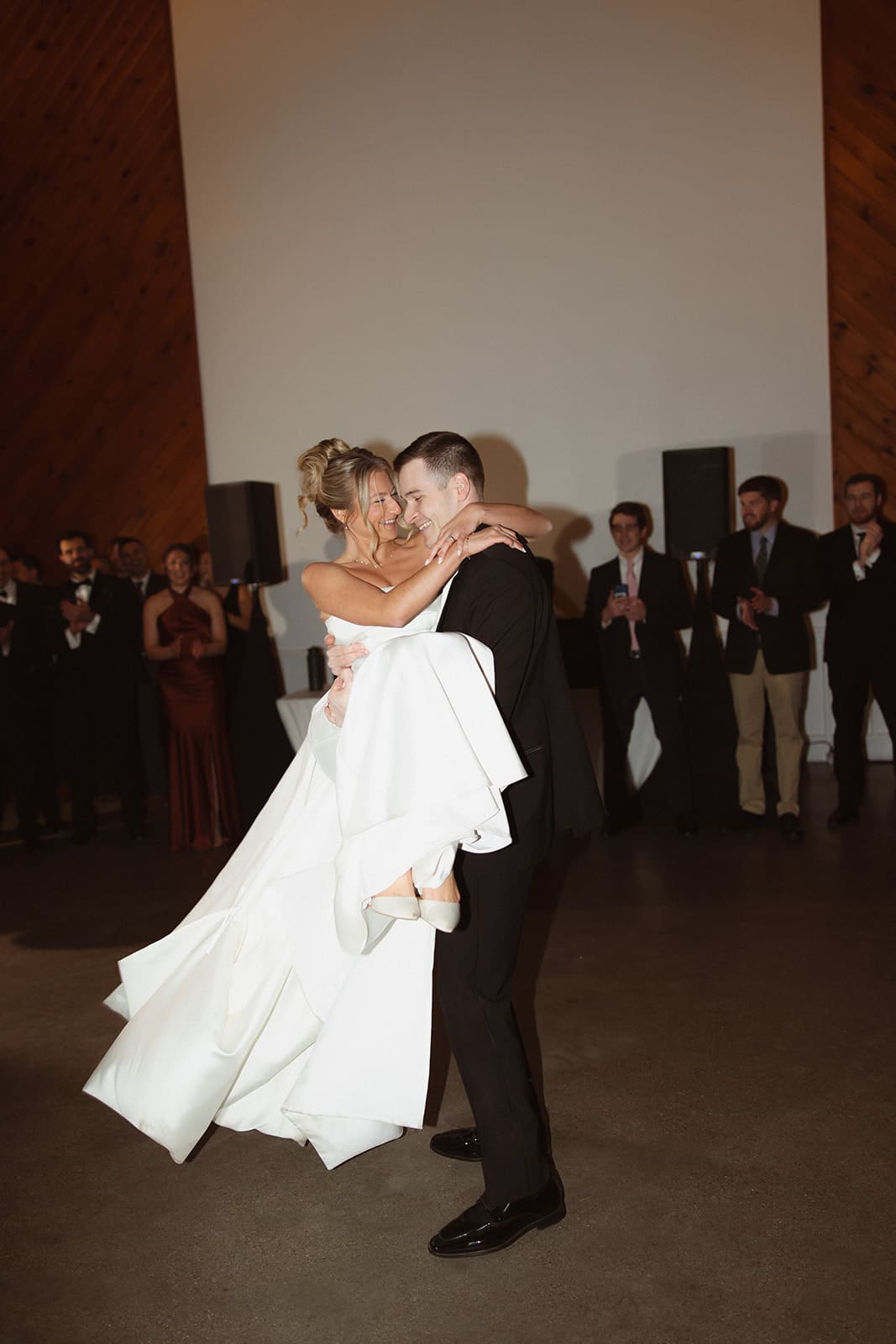 Groom lifting bride during joyful reception dance surrounded by cheering guests.