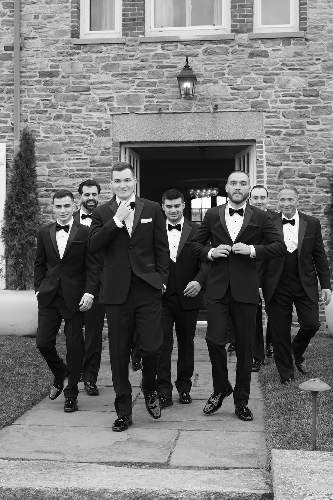 Groom walking confidently with groomsmen in black tuxedos outside historic stone estate.