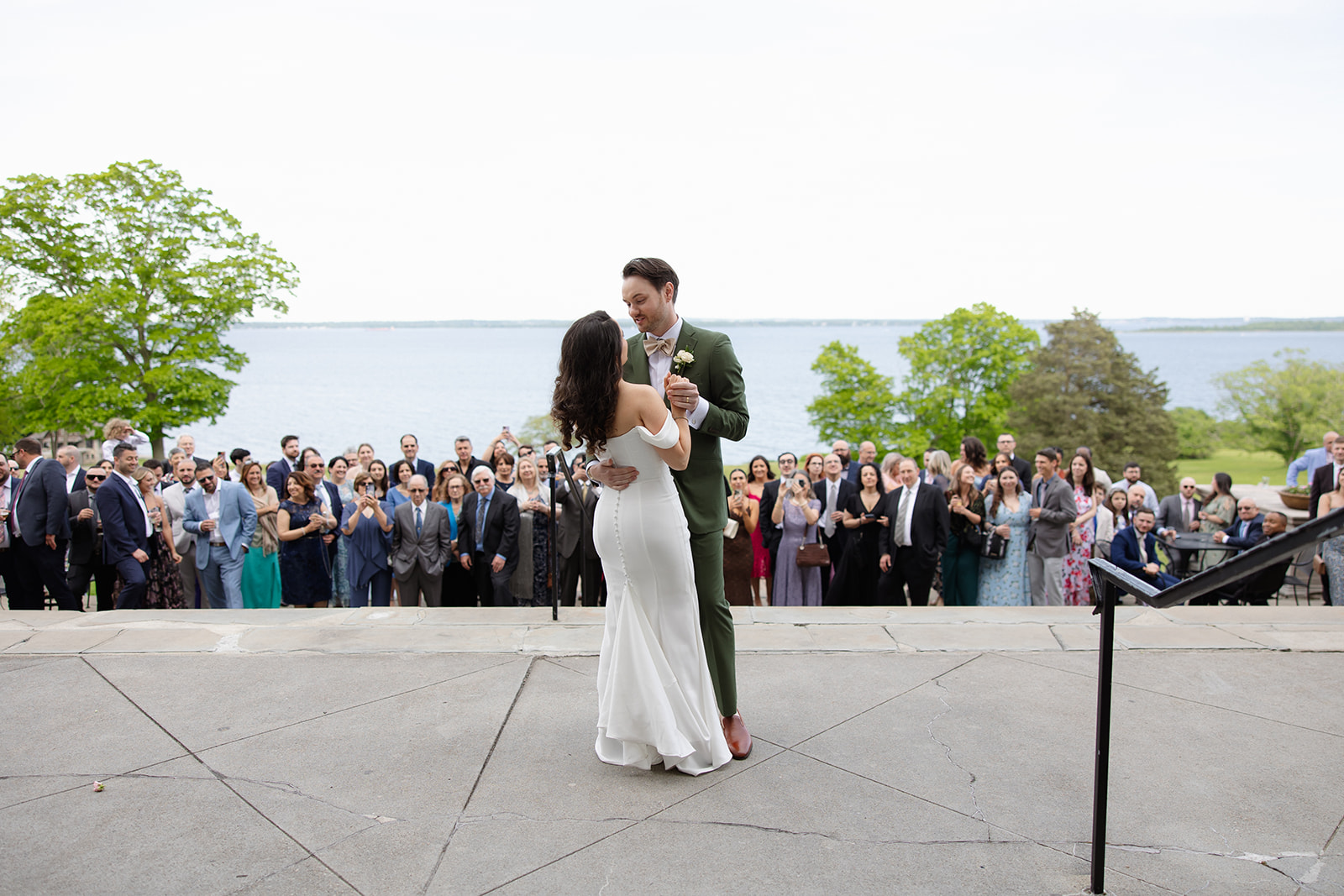 The couple shares their first dance outdoors with guests gathered closely around them overlooking the water.
