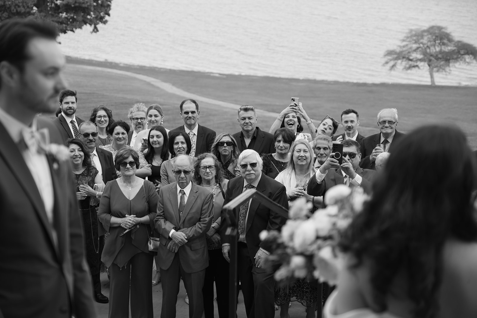 Guests watch the ceremony with quiet attention, some capturing the moment on their phones in a black and white frame.
