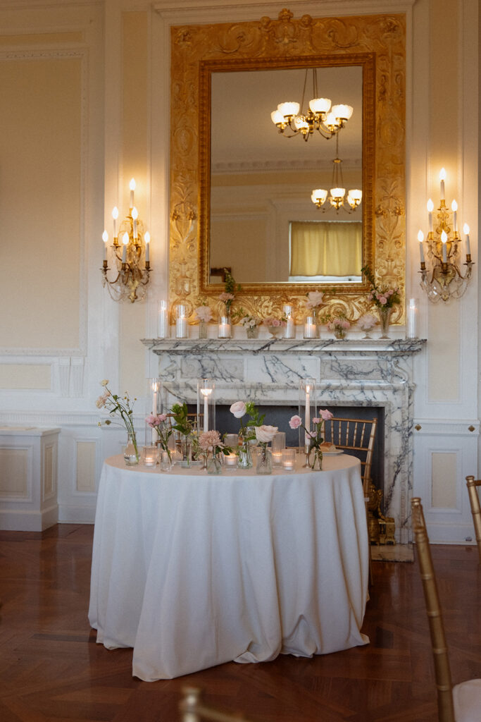 A softly styled table with candles and floral arrangements sits in front of a marble fireplace and gilded mirror.
