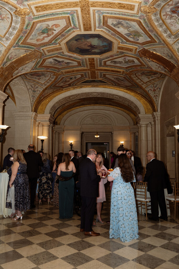 Guests gather for cocktail hour beneath an ornate painted ceiling in a historic venue during a colorful wedding.
