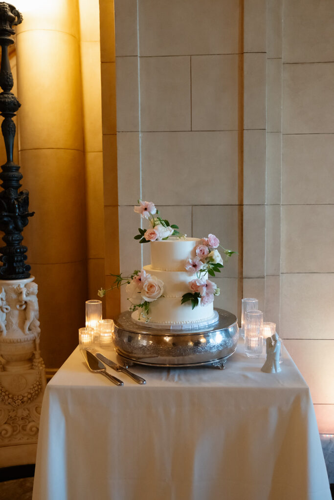 A simple two-tier wedding cake with soft floral details sits surrounded by candlelight during a colorful wedding reception.
