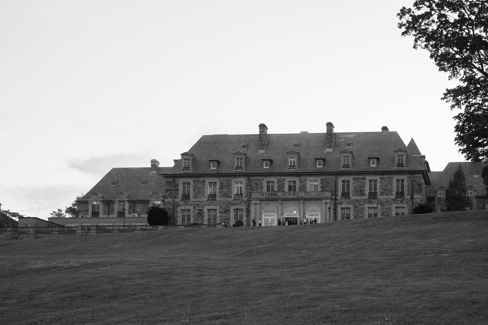 A wide black and white view of a grand estate set on a hill, offering a quiet sense of place before the celebration begins.
