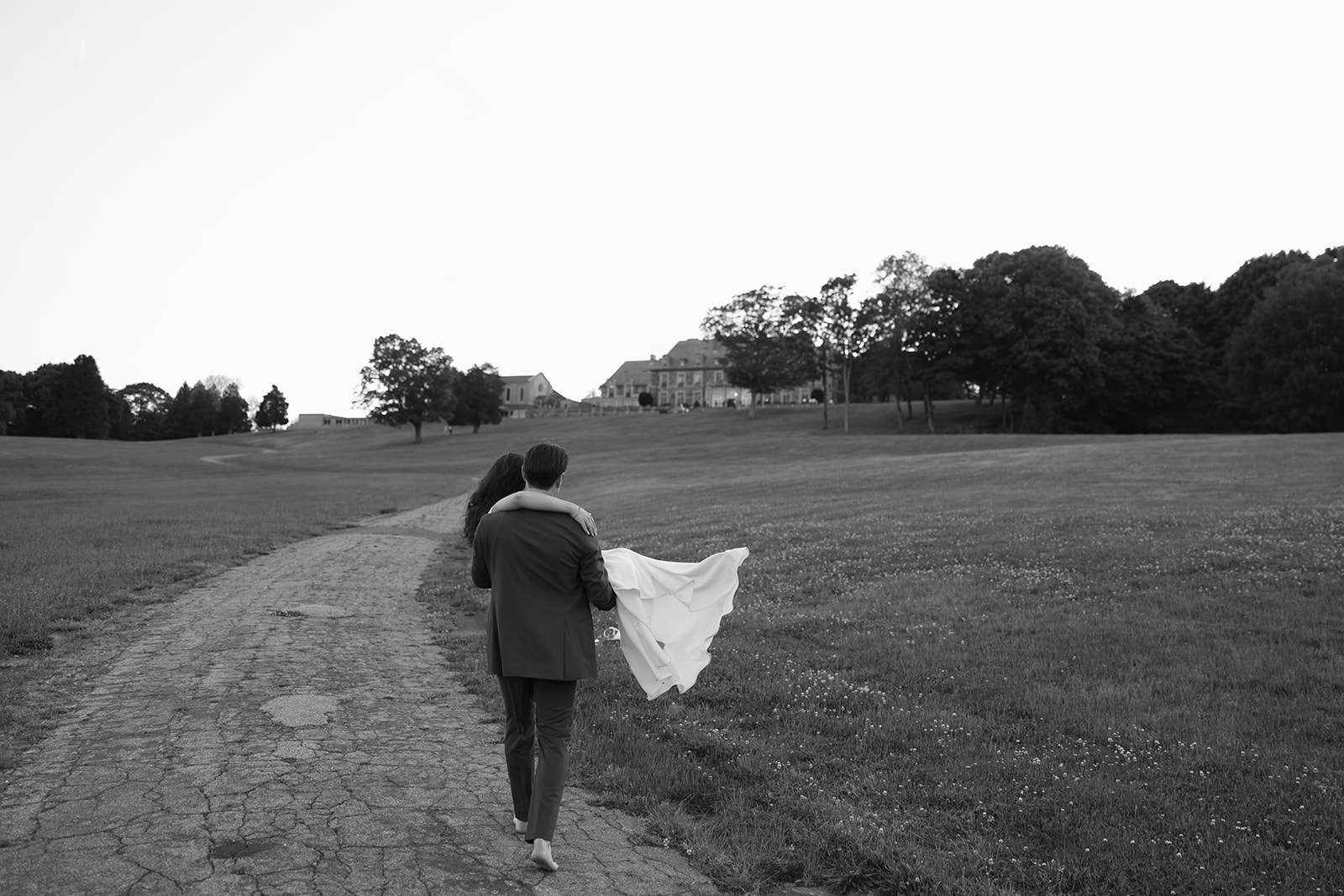 The couple walks away together along a quiet path at dusk, the estate visible in the distance in a black and white frame.
