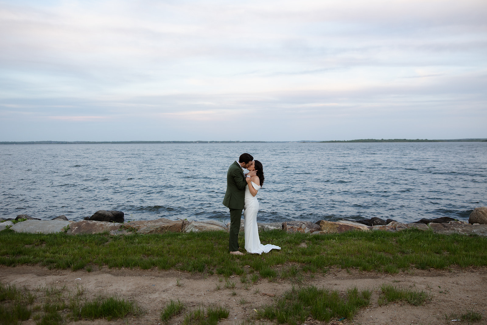 The couple stands close along the shoreline, the water stretching behind them in a calm, open landscape.
