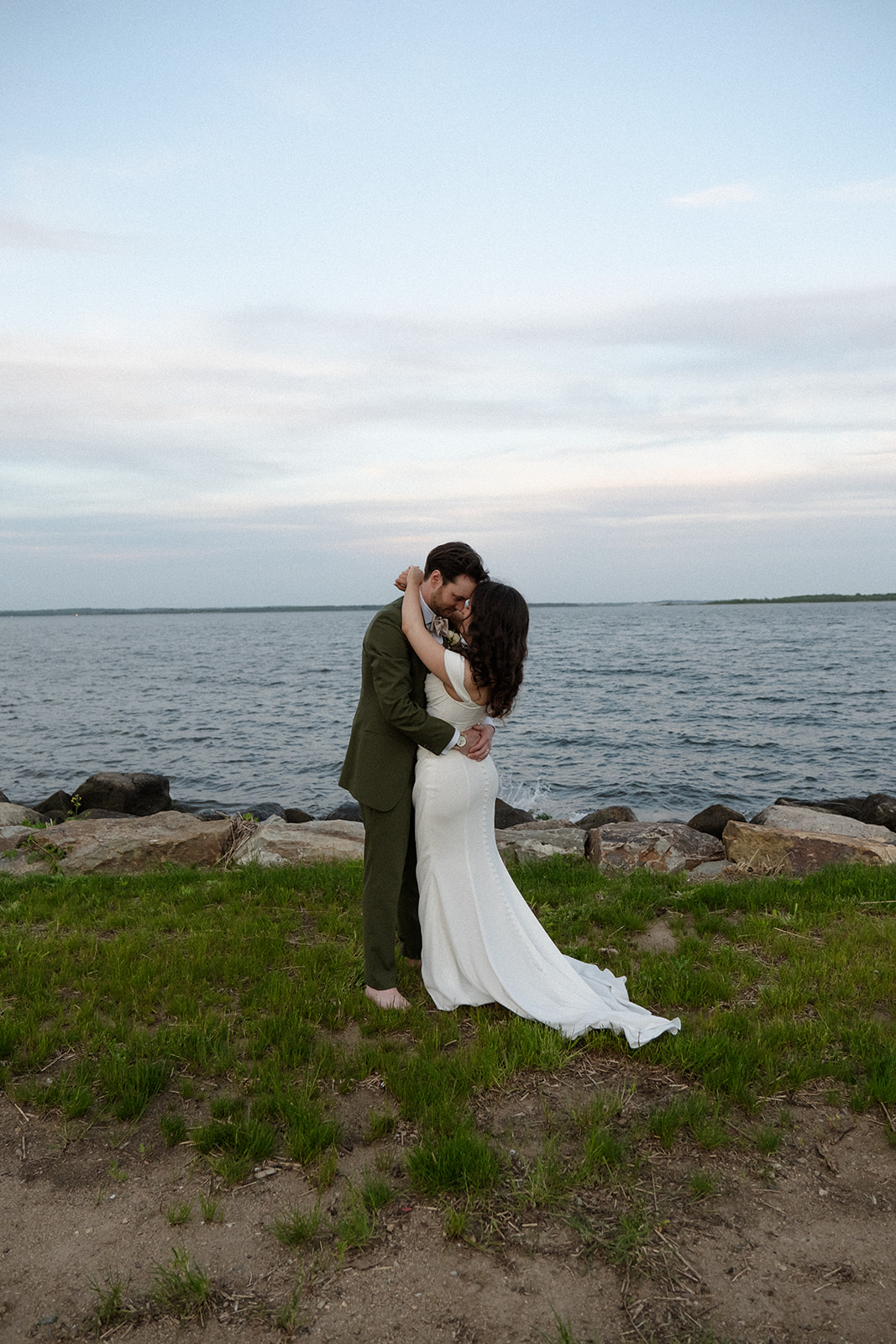 The couple stands close along the shoreline, wrapped in each other during a colorful wedding portrait by the water.
