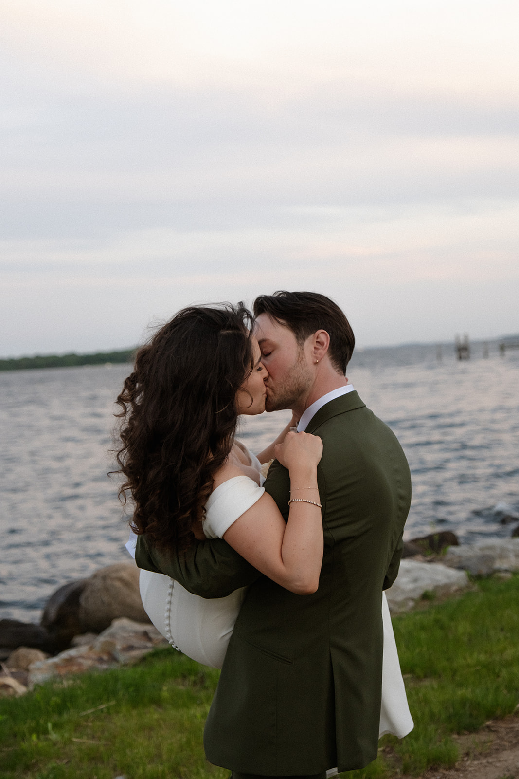 A close, intimate kiss by the water as the bride’s dress moves softly in the evening light.
