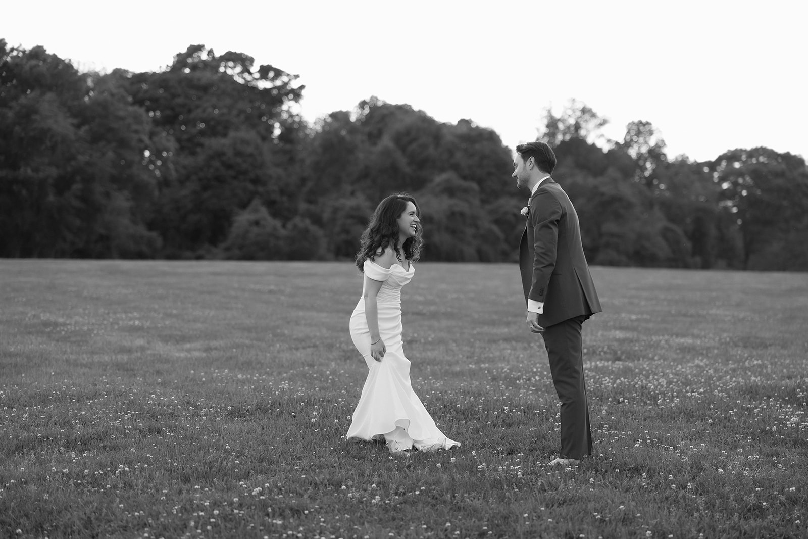 The bride and groom laugh together in an open field, a quiet and candid moment in black and white.
