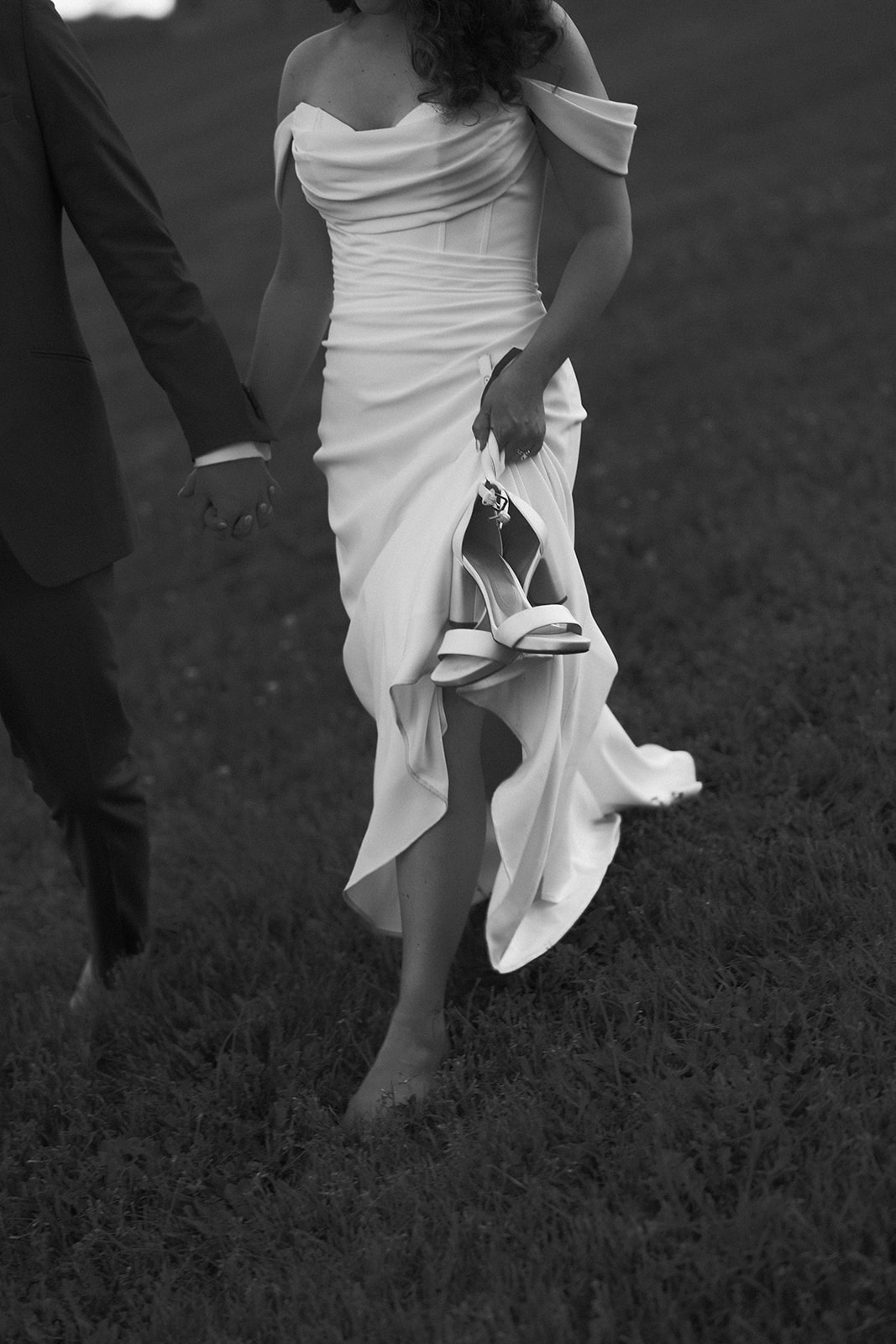 The bride walks barefoot through the grass holding her shoes, her dress gathered gently in a black and white frame.
