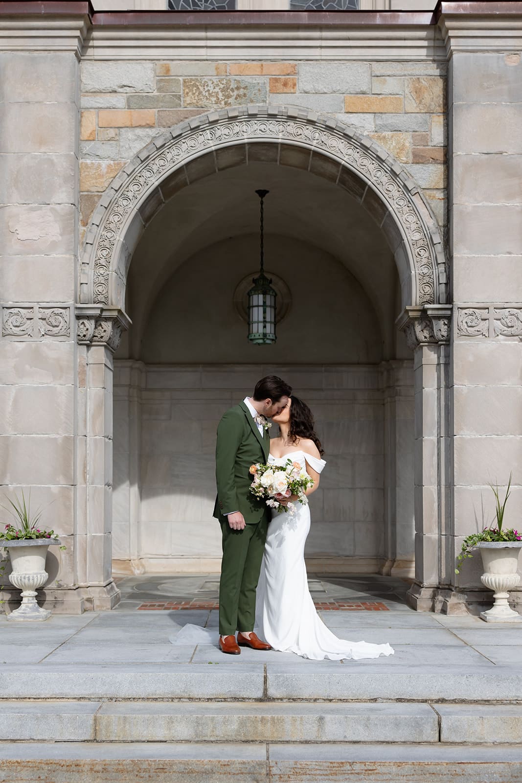 The bride and groom stand close beneath a stone archway, holding a soft garden bouquet during a colorful wedding day.
