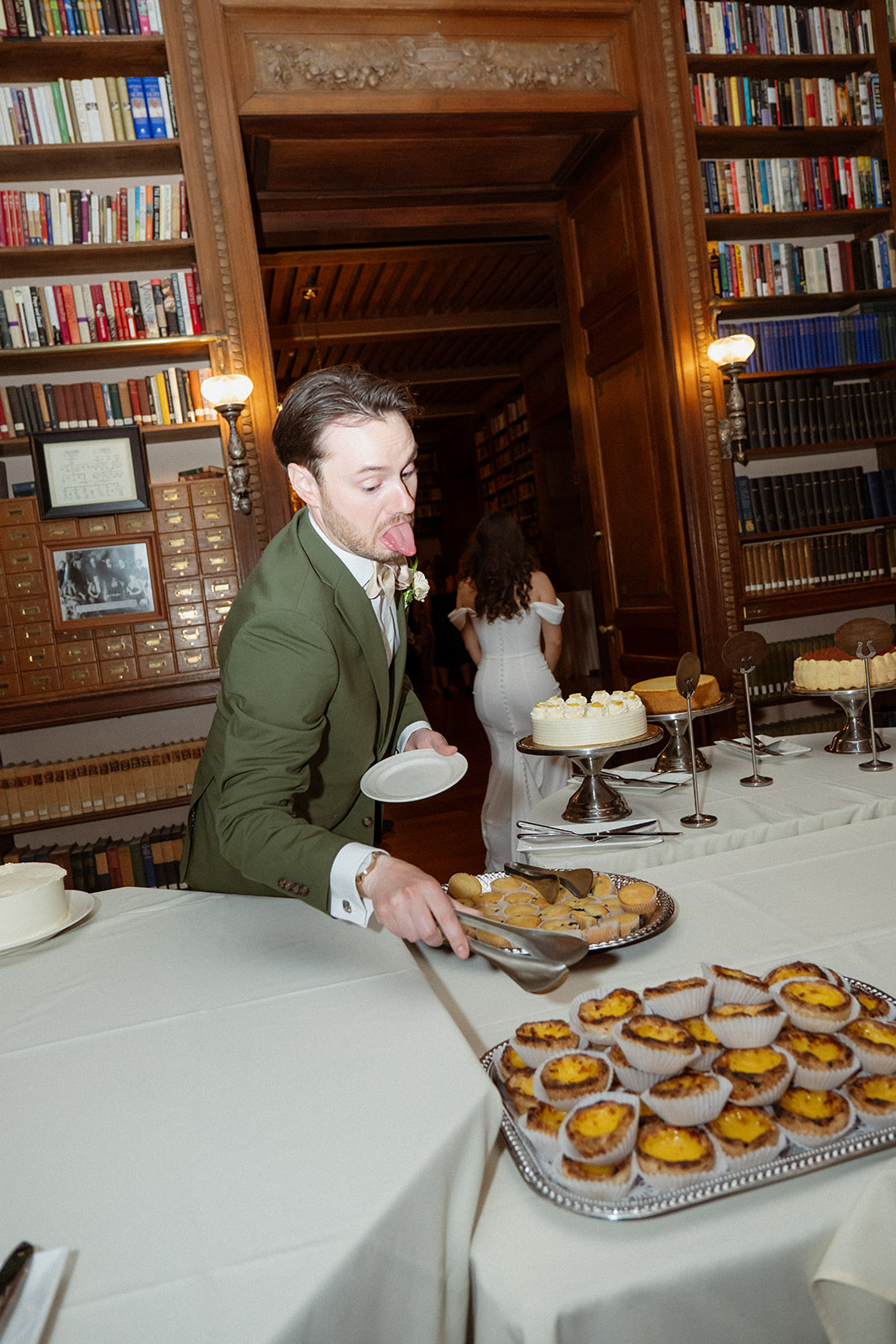 The groom reaches for pastries at the dessert table while guests mingle nearby during a colorful wedding reception.
