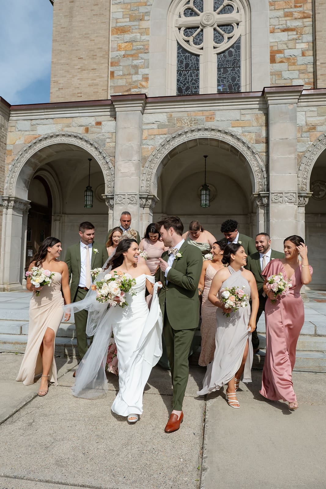 The couple walks out with their wedding party beneath the stone arches, surrounded by soft tones and movement.
