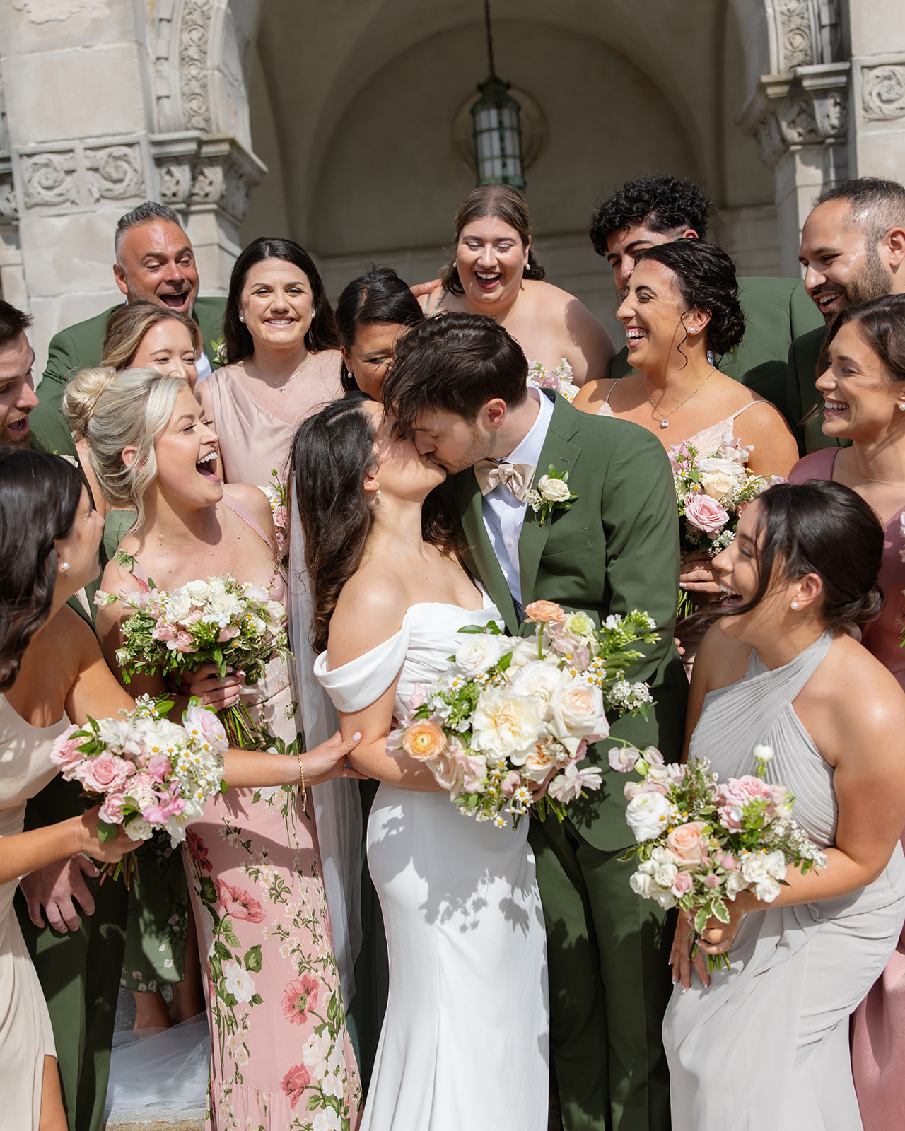 The couple is surrounded by their wedding party, sharing a kiss while everyone reacts with joy during a colorful wedding celebration.
