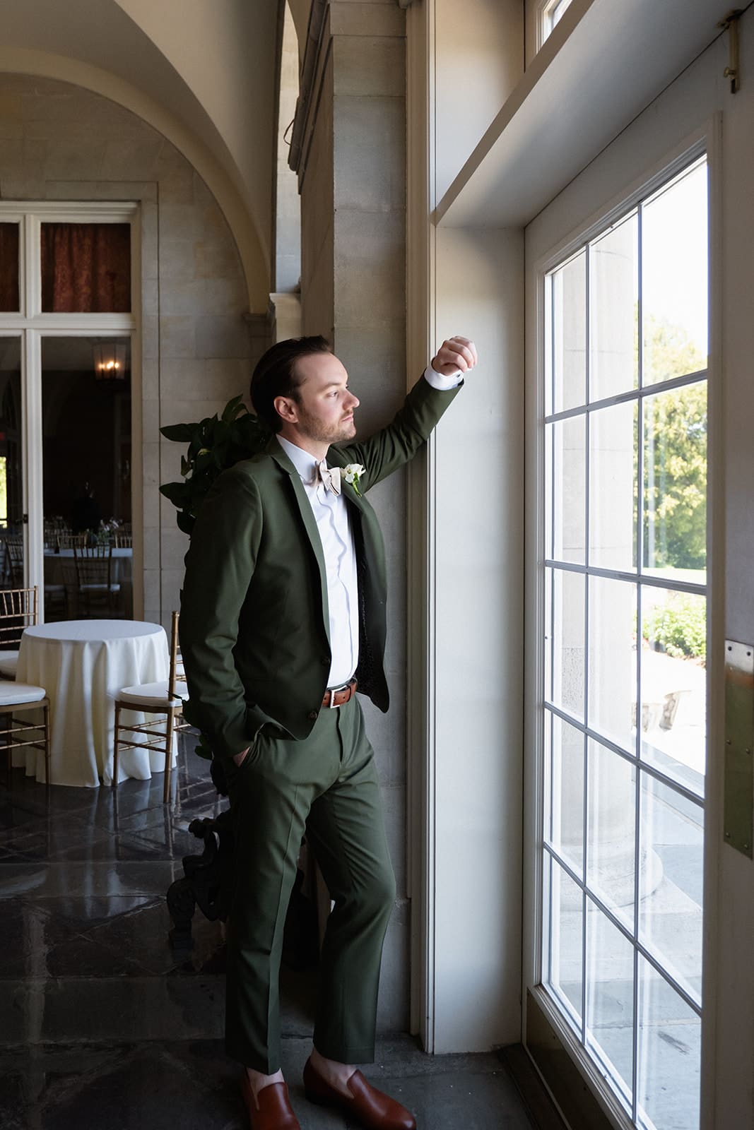 The groom stands by the window in soft natural light, taking in a quiet moment before the ceremony.
