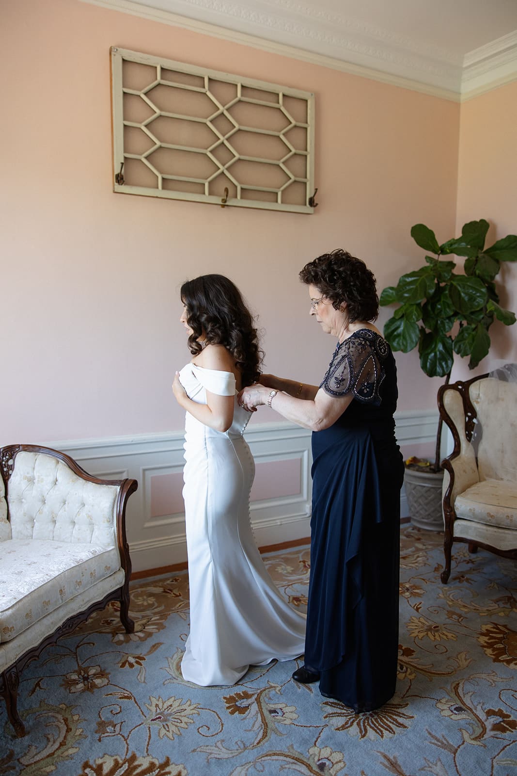 A mother helps the bride into her dress in a softly lit room, a quiet and meaningful getting-ready moment.
