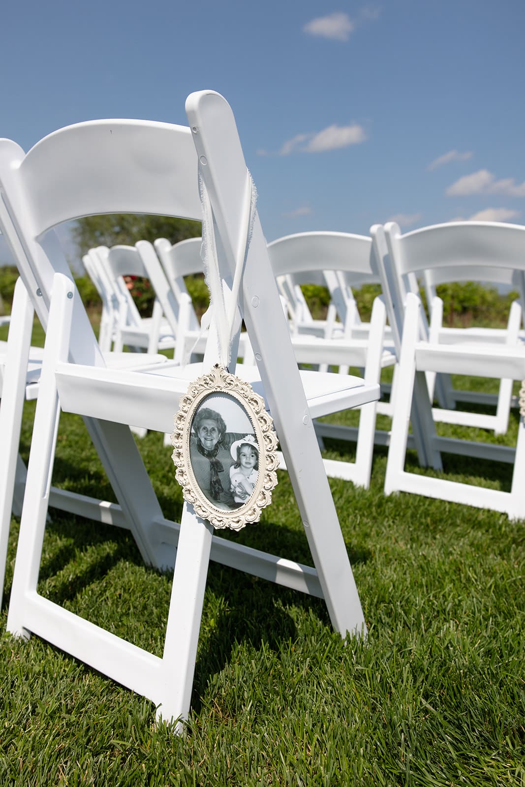 Framed photo attached to a white ceremony chair at an outdoor wedding, honoring a loved one during the ceremony.