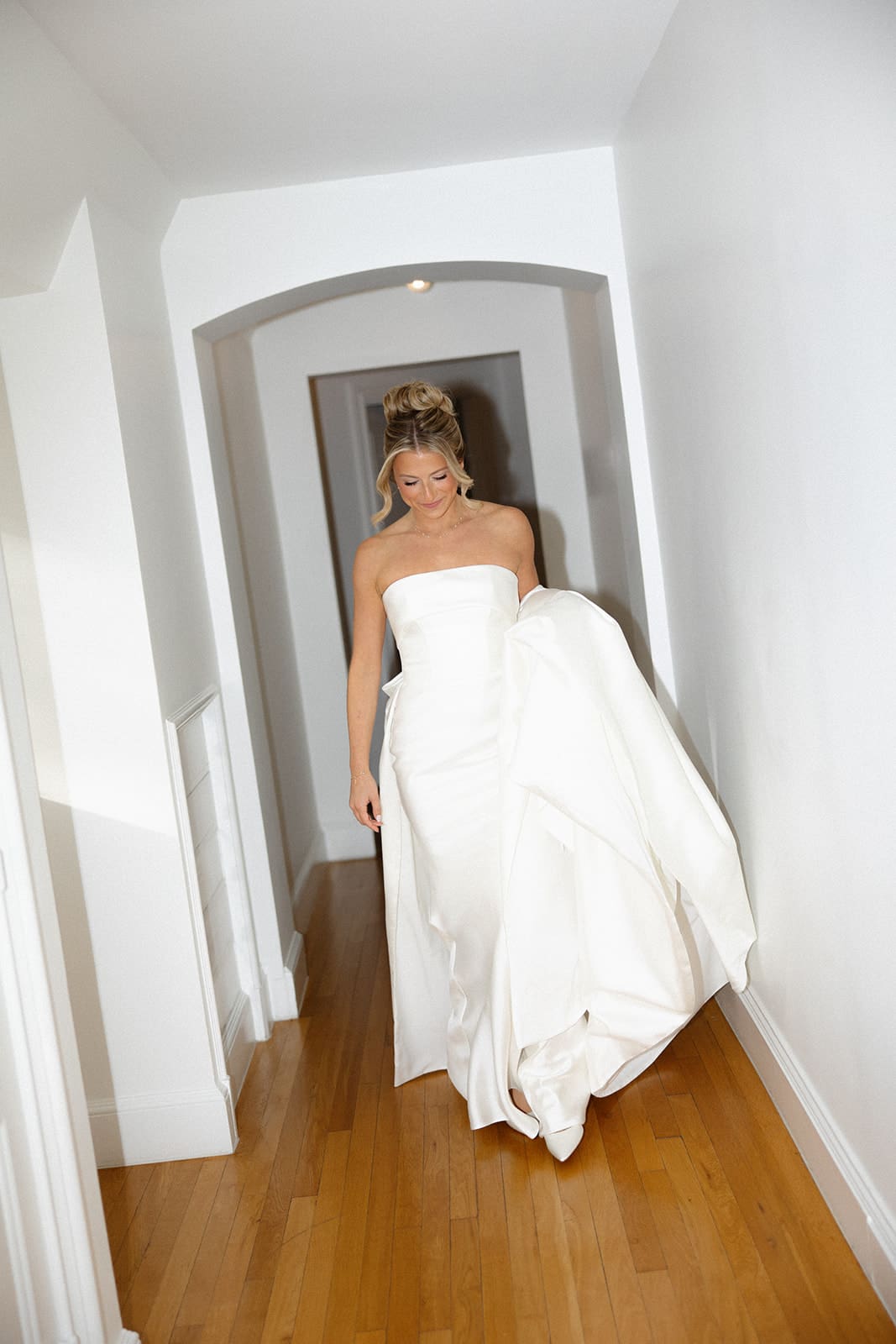 Bride walking down a hallway holding the skirt of her modern strapless wedding dress on the morning of her wedding.
