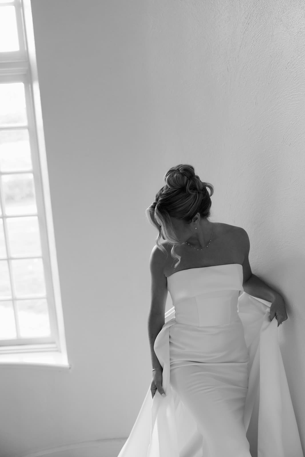 Black and white bridal portrait by a tall window highlighting a minimalist strapless wedding dress.
