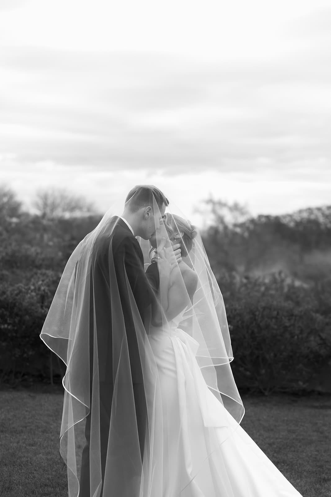 Black and white portrait of bride and groom sharing a quiet moment beneath the veil outdoors on their wedding day.
