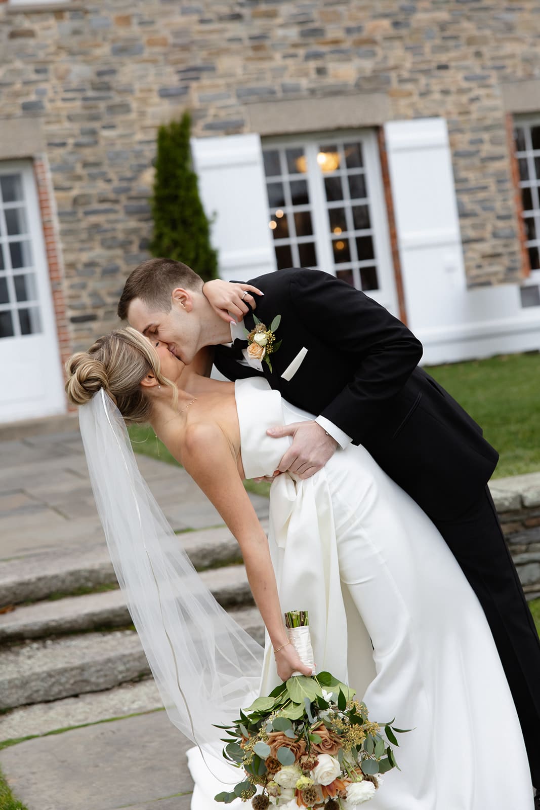 Groom dipping the bride for a kiss while holding her bouquet in front of a historic stone wedding venue.
