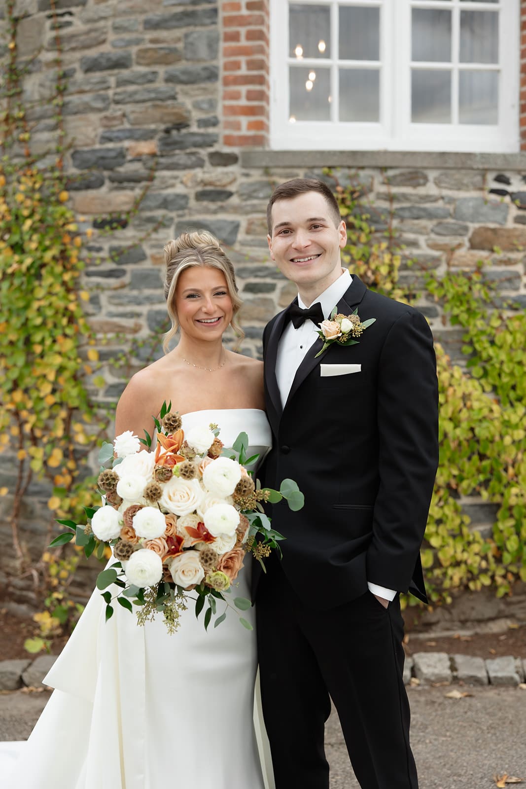 Bride and groom posing with a fall-toned bouquet in front of a historic stone estate, a classic setting among Wedding Venues in Rhode Island.
