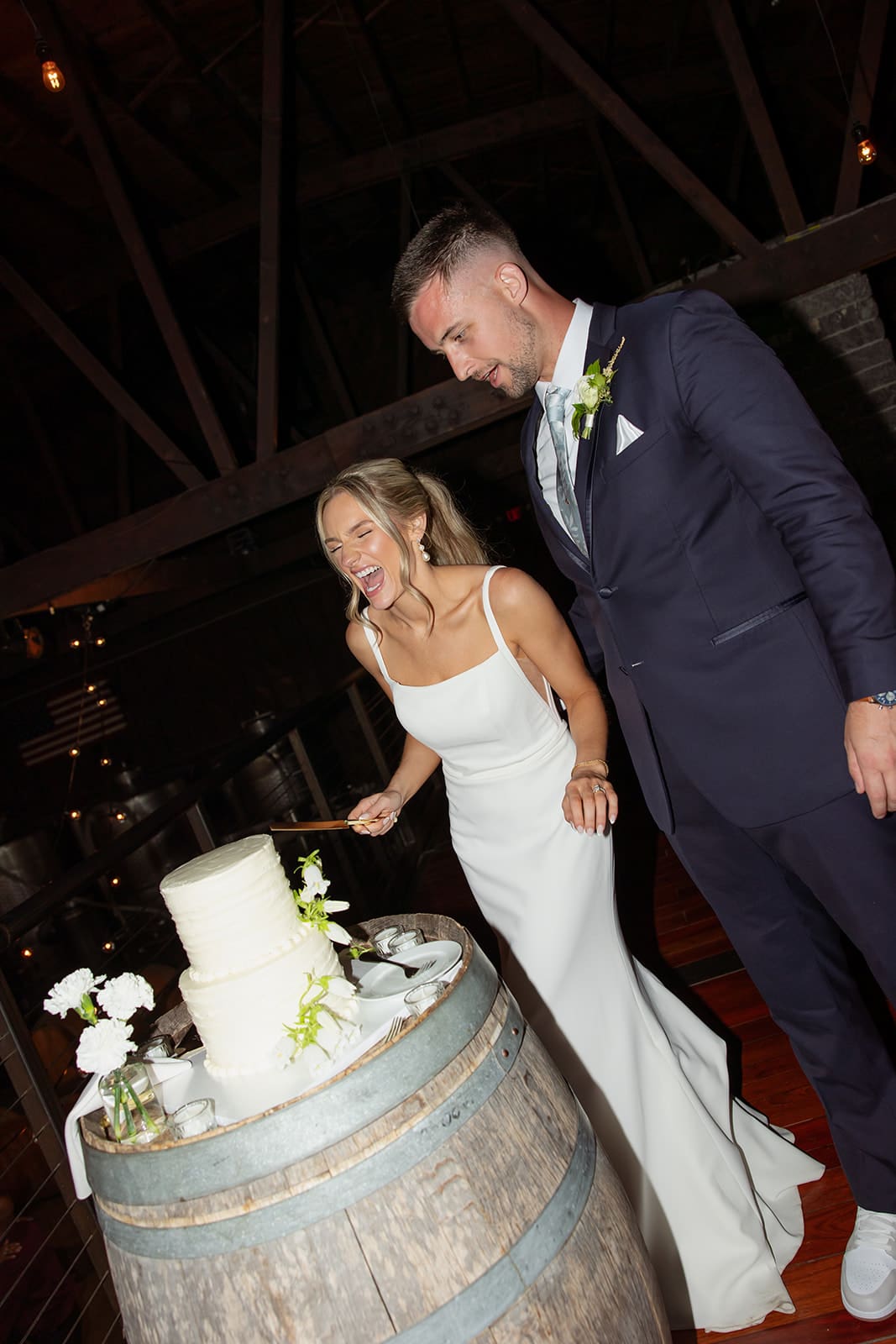 Bride and groom smiling as they cut their wedding cake displayed on a wooden barrel during their barn reception.