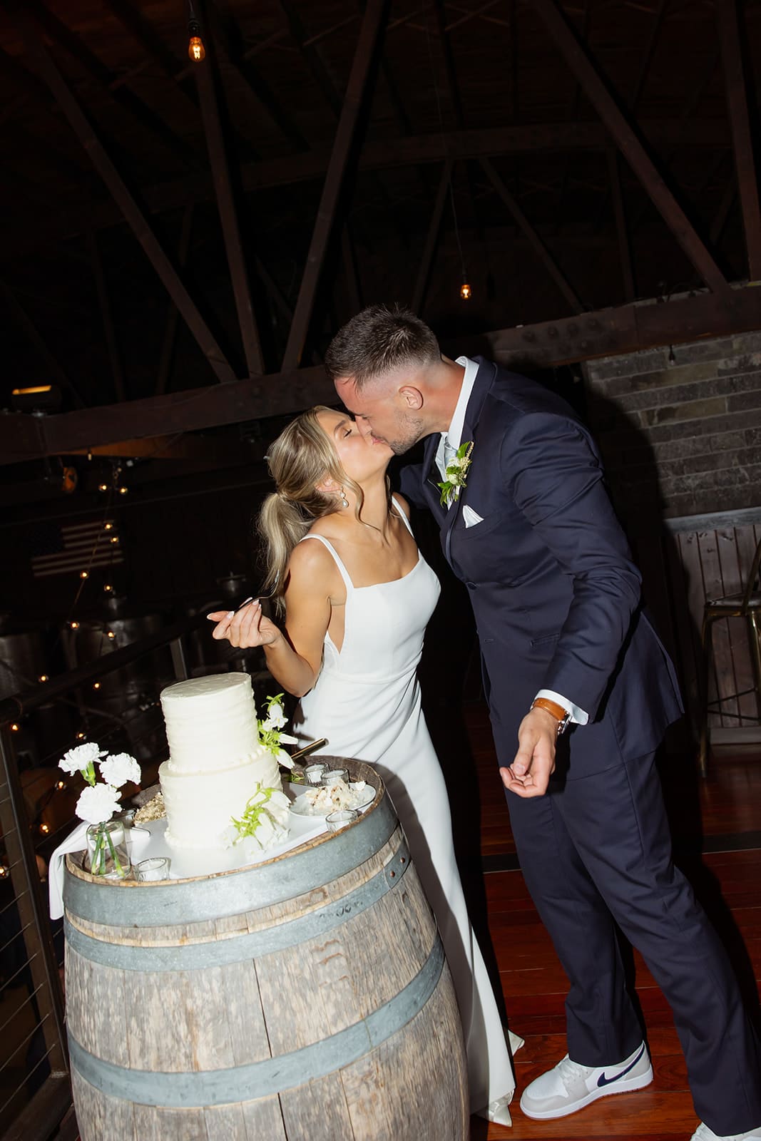 Bride and groom sharing a kiss while cutting their wedding cake during their rustic indoor reception.