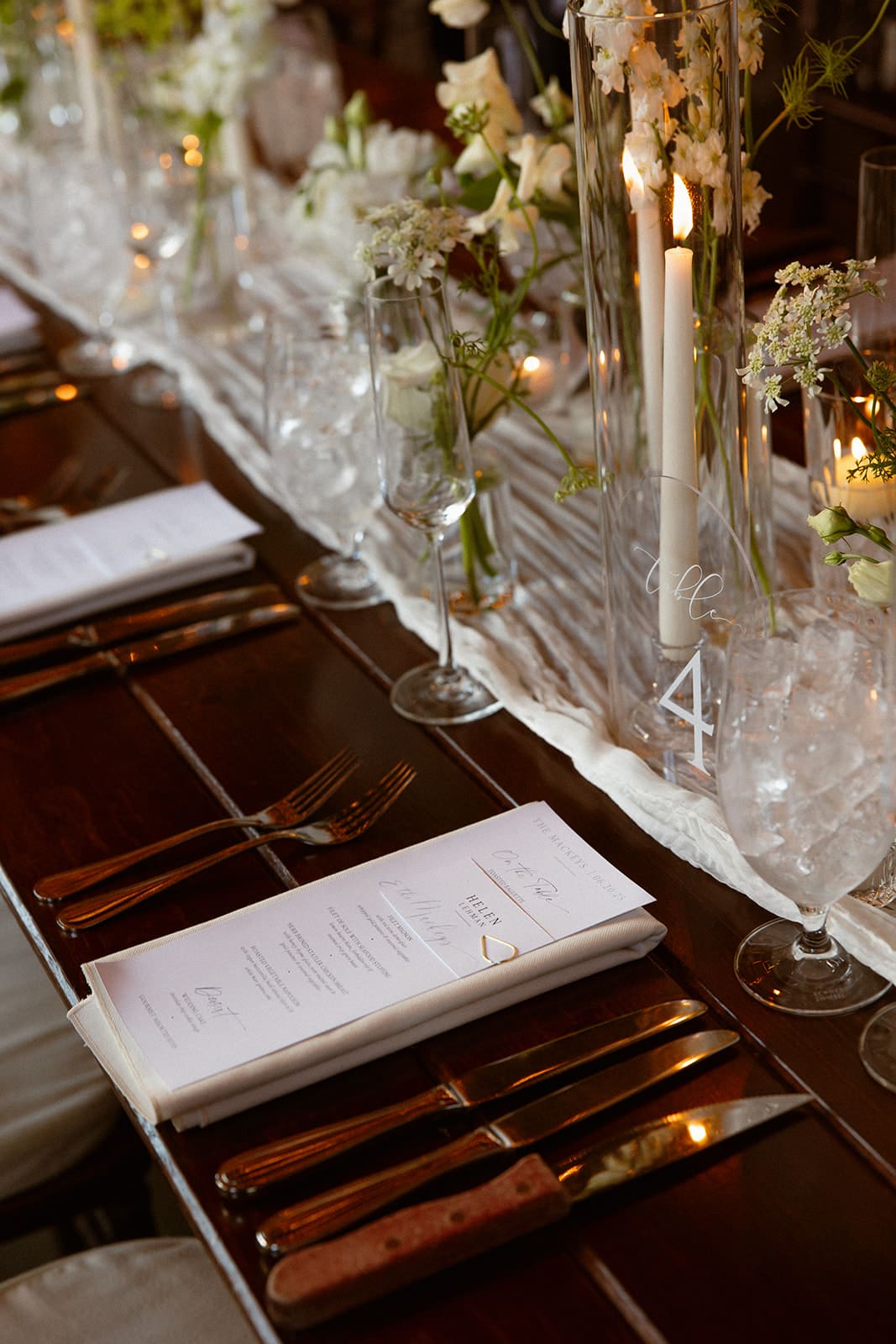 Elegant wedding reception table with candles, floral centerpieces in glass vases, and place settings with menus on a long wooden table.