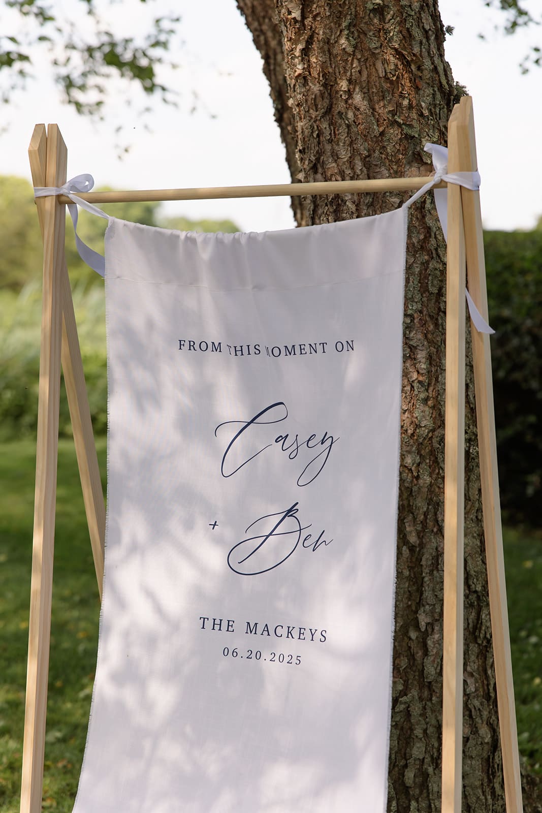 Custom wedding welcome sign with couple’s names displayed outdoors beneath a tree
