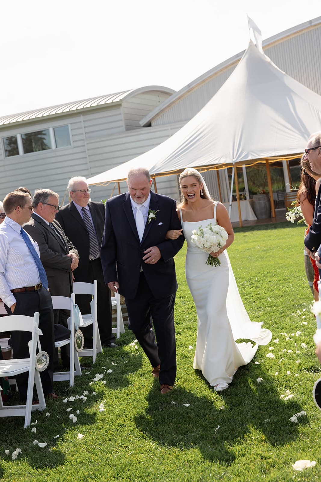 Bride walking down the aisle with her father during an outdoor wedding ceremony on a sunny day, with guests seated on white chairs under a white tent.
