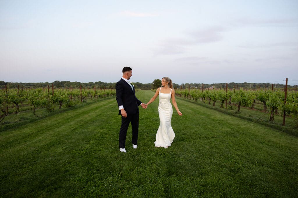 Newlyweds walking hand in hand through vineyard rows at sunset
