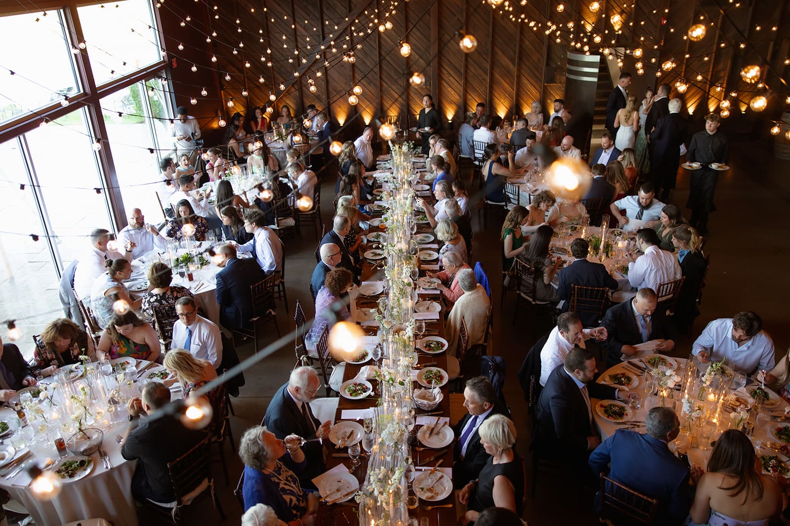 Guests enjoying dinner beneath string lights inside a modern barn Connecticut wedding venue
