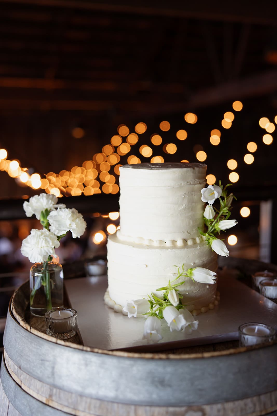 Two-tier white wedding cake decorated with small white flowers displayed on a wooden barrel with warm string lights in the background.
