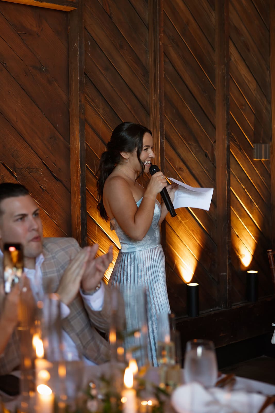 Bridesmaid giving a heartfelt speech during a candlelit wedding reception in a rustic barn setting.