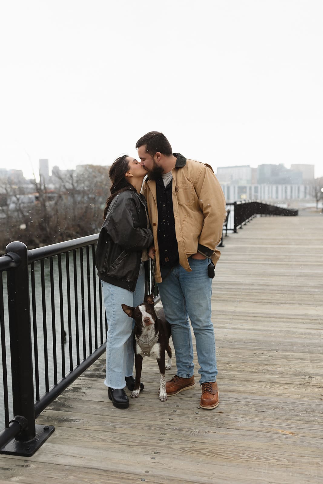 Couple kissing on a wooden boardwalk beside their dog with the waterfront city skyline in the background.
