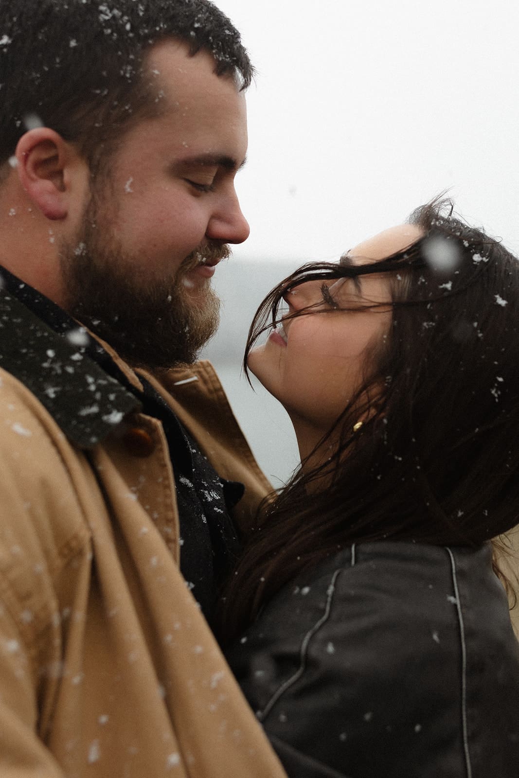 Romantic snowy close-up of a couple about to kiss during a winter engagement session by the water.
