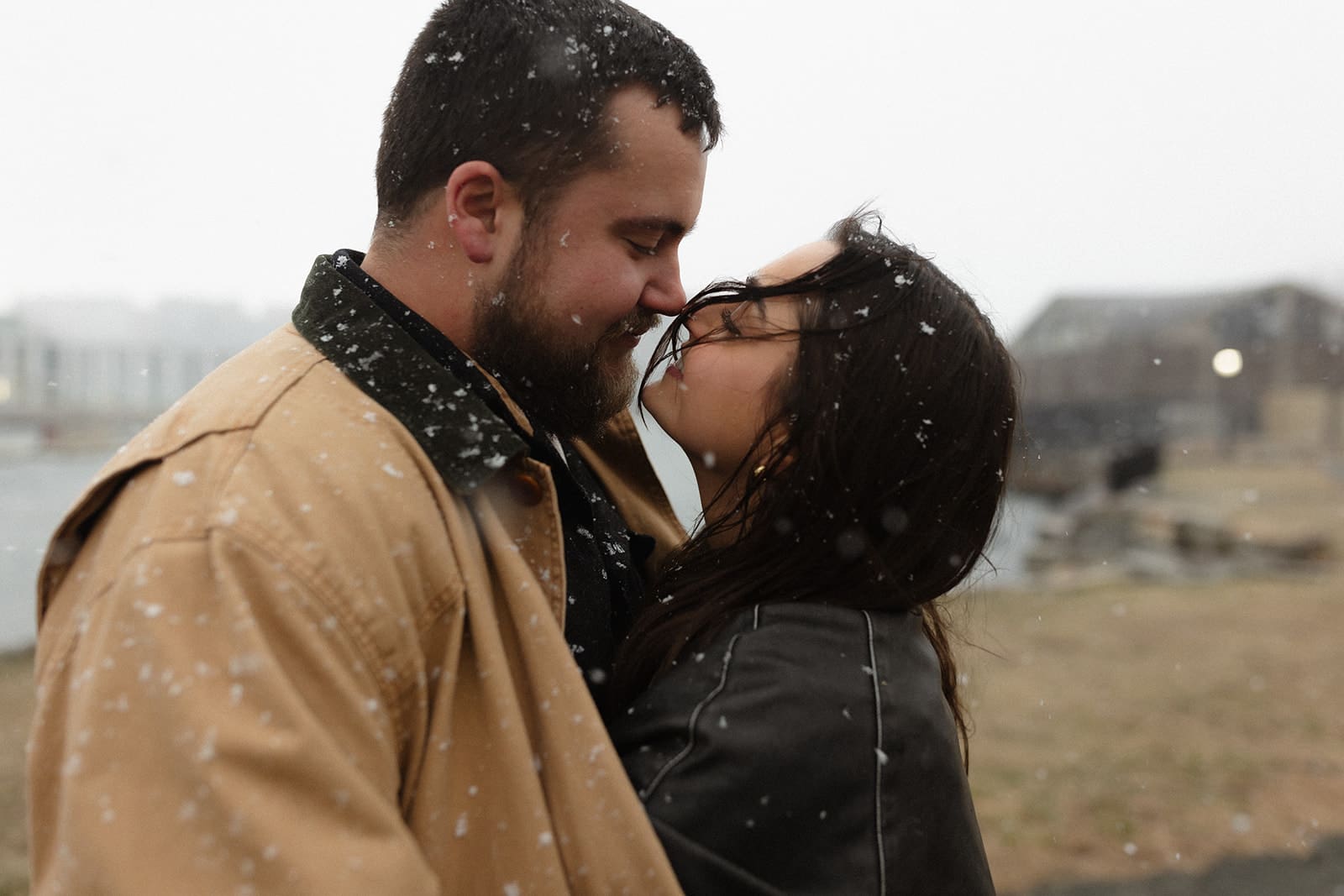 Romantic close-up of a couple embracing in falling snow along the waterfront during a winter engagement session.
