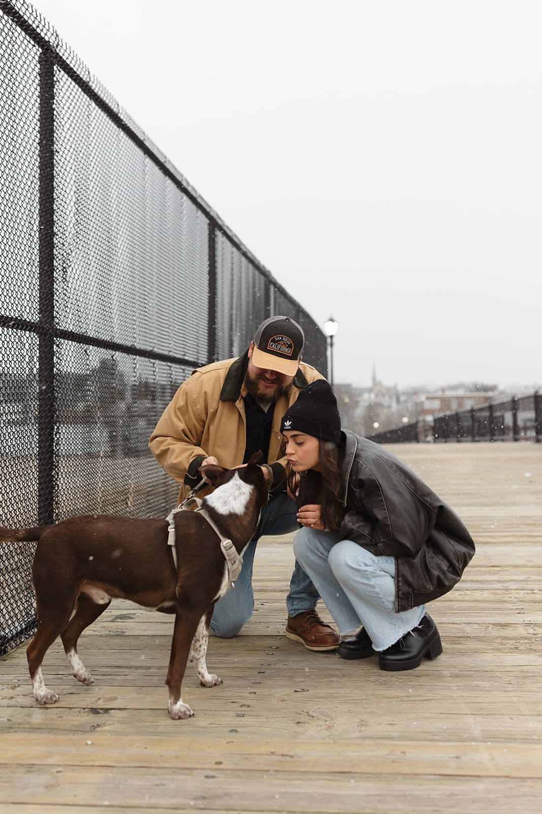 Couple crouching to pet their dog on a wooden boardwalk during a candid winter engagement session.
