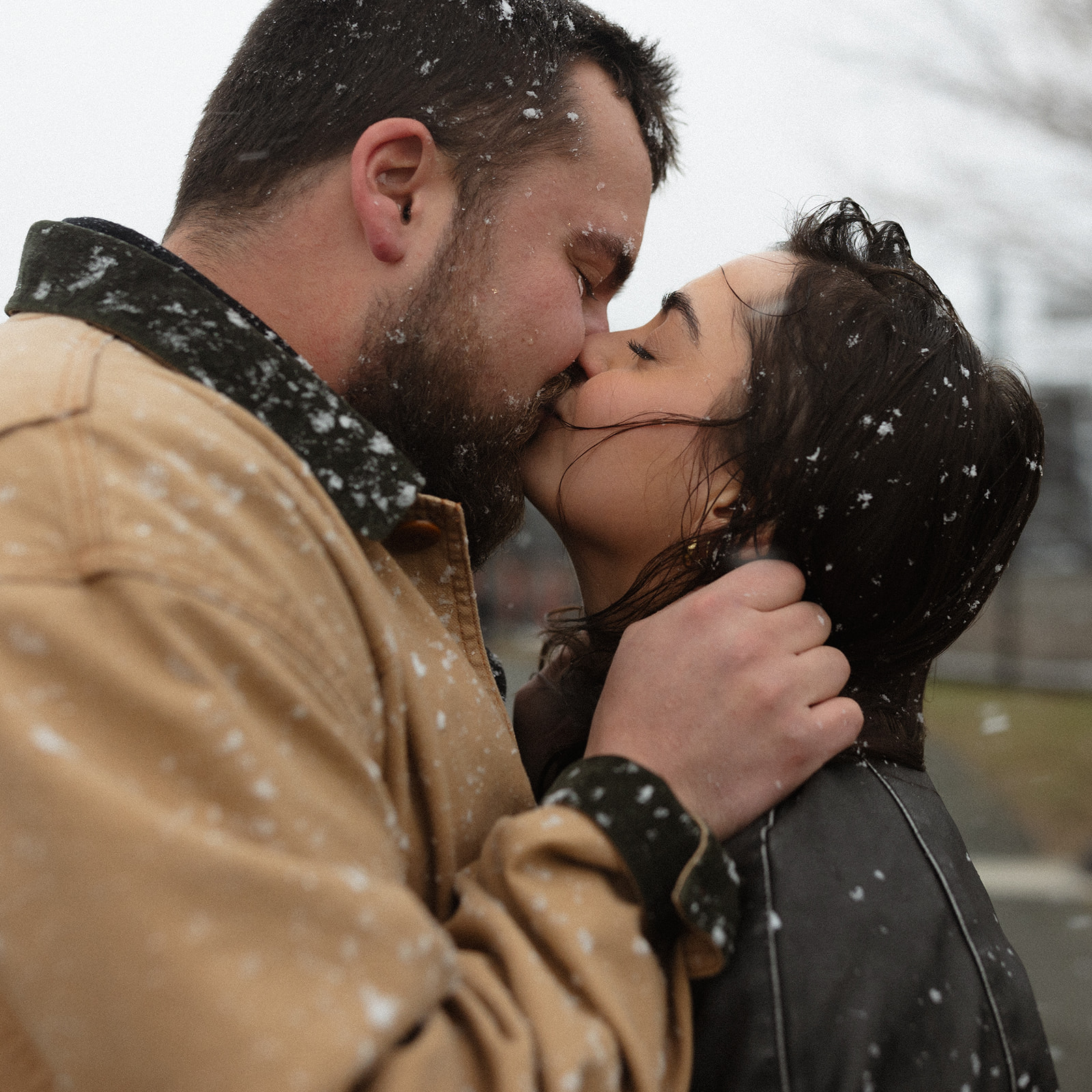 Snowy kiss between a couple wearing cozy winter engagement session outfits including a tan jacket and leather jacket.
