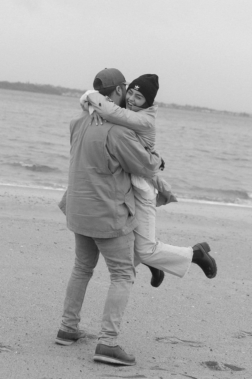 Playful moment as a couple spins and hugs along the shoreline during a breezy coastal engagement session.
