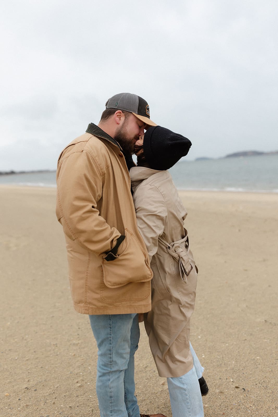 Tender beach portrait of a couple embracing in neutral winter engagement session outfits on a cloudy coastal day.
