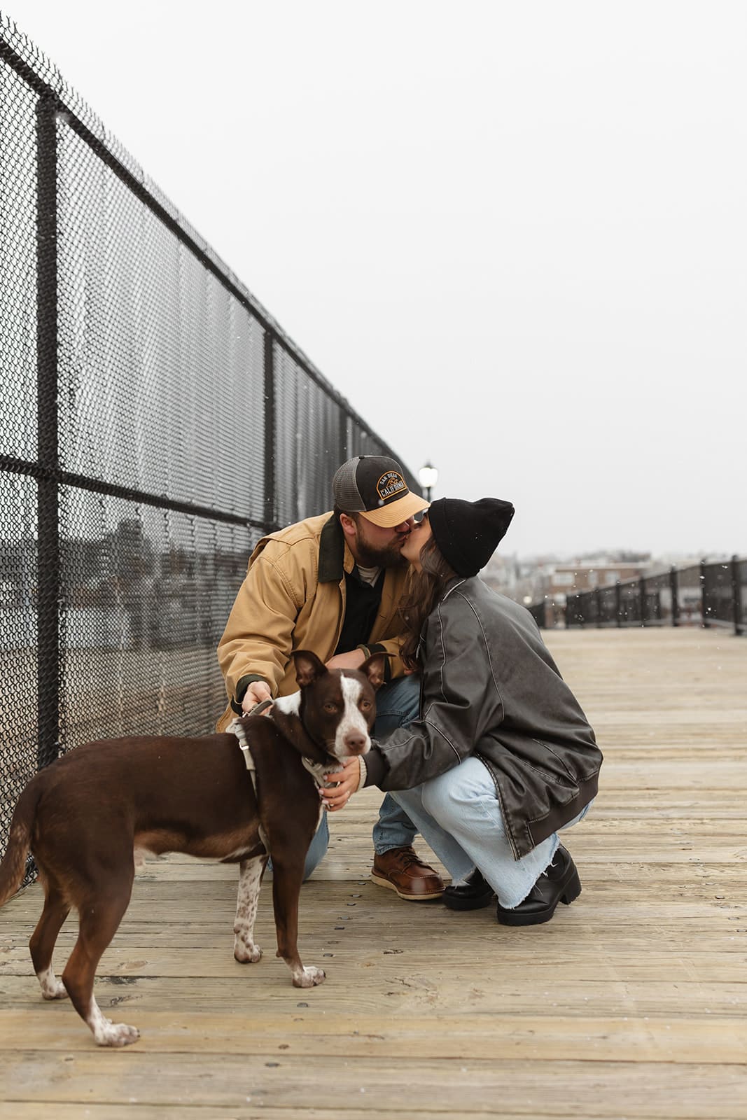 Couple kissing beside their dog on a wooden boardwalk during a candid coastal engagement session.
