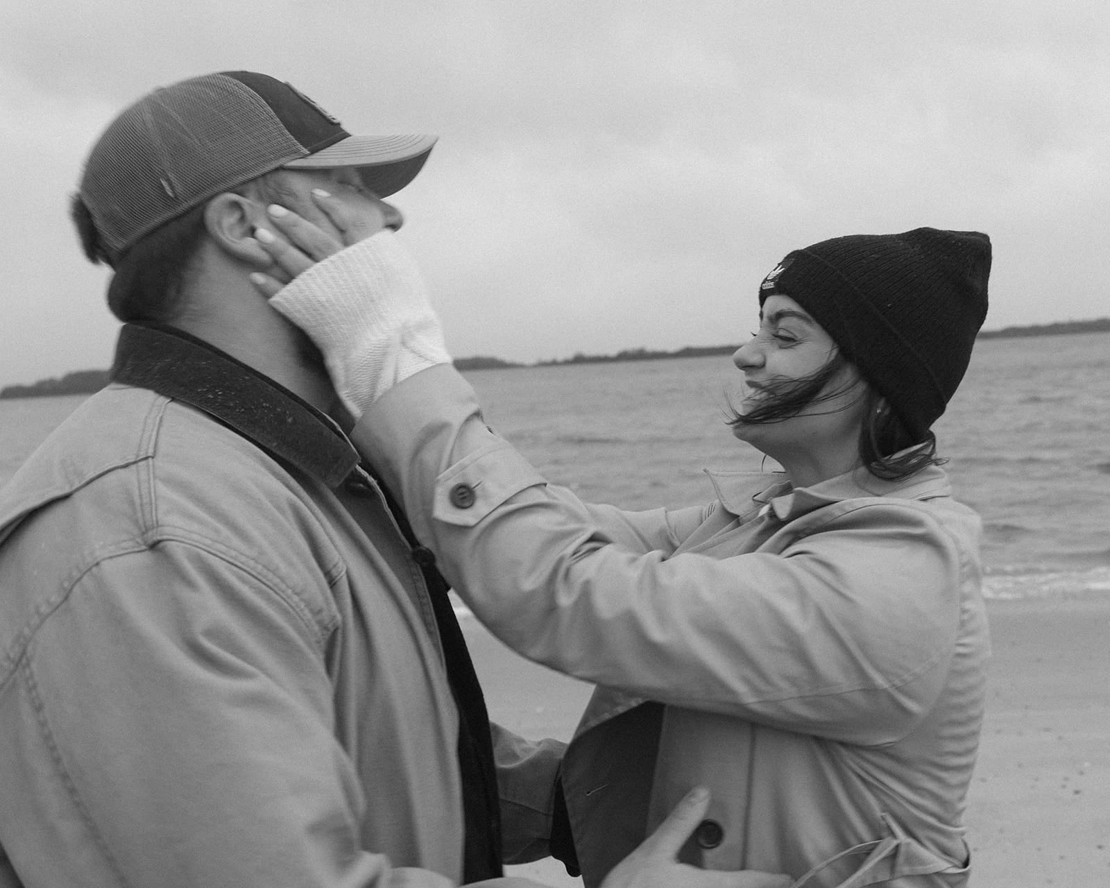 Windy black and white engagement portrait as a couple laughs together by the ocean in warm winter engagement session outfits.
