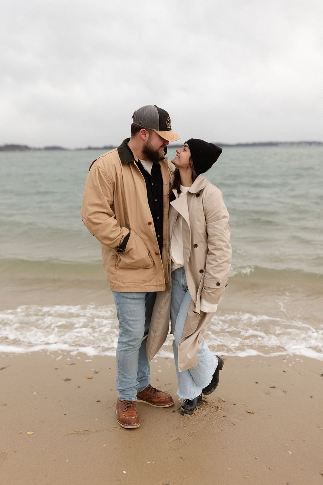 Cozy beach portrait of a smiling couple bundled in warm winter engagement session outfits beside the ocean.
