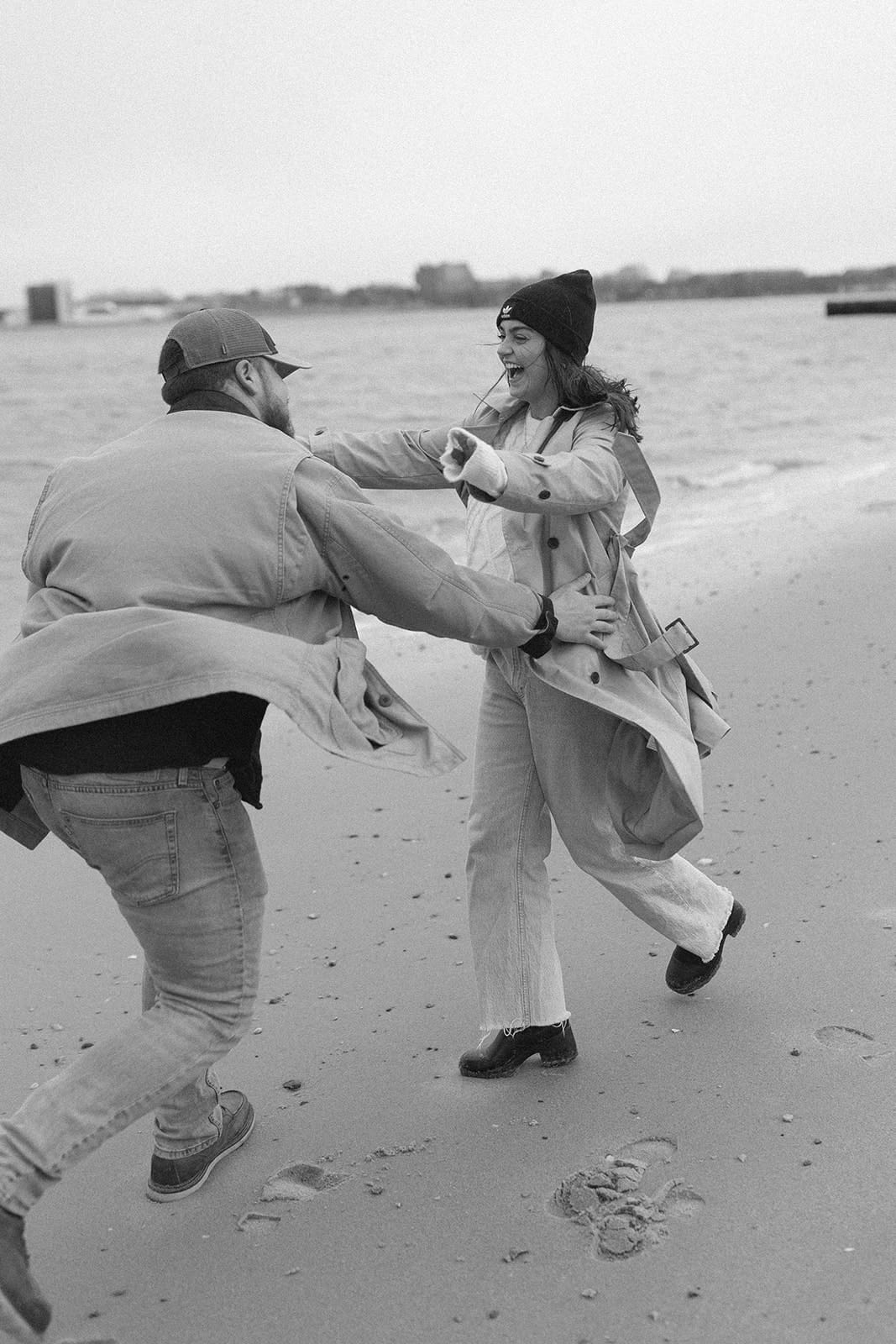 Joyful moment as a couple spins together on the shoreline wearing layered winter engagement session outfits for a breezy coastal session.
