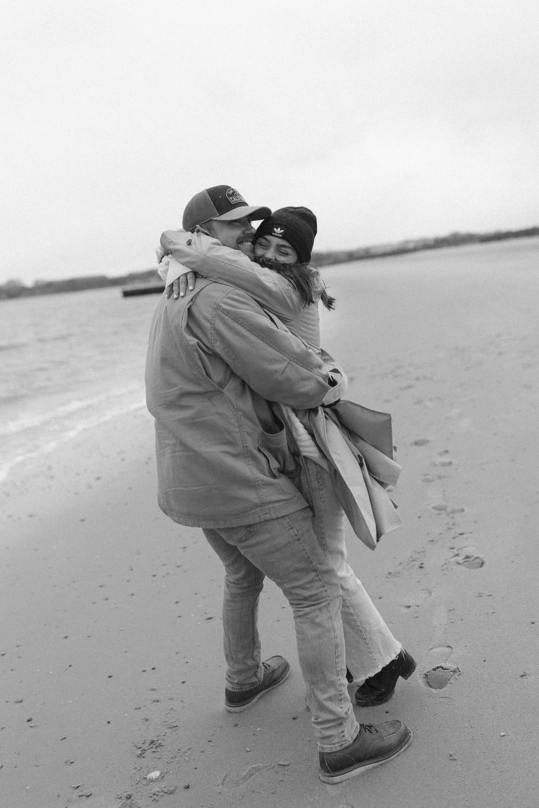Playful black and white engagement photo of a couple hugging while walking along a quiet beach.
